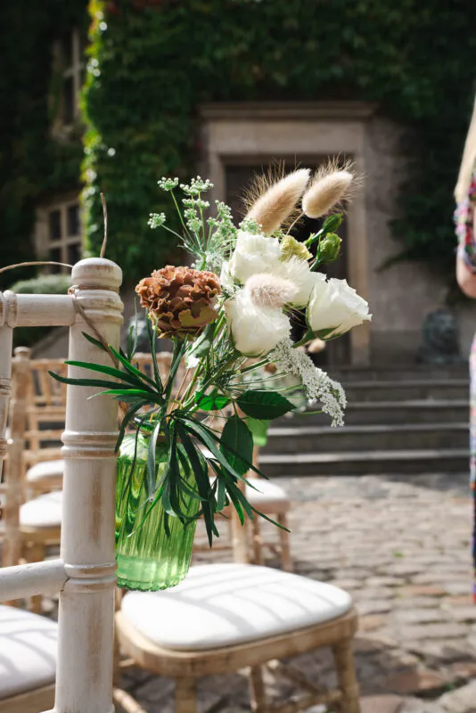 A rustic wedding chair aisle end is decorated with a vibrant green glass bottle holding a floral arrangement of white roses, dried grasses, and green foliage against an outdoor stone courtyard backdrop.