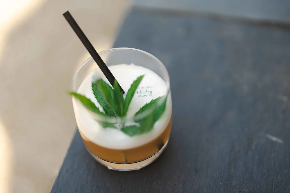 A refreshing wedding cocktail garnished with fresh mint leaves and a personalised foam topper sits on a dark slate surface during the drinks reception.
