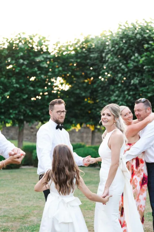 A bride in a sleek sleeveless dress and a groom in a white shirt and bow tie happily hold hands and dance outdoors in a garden with family and guests.