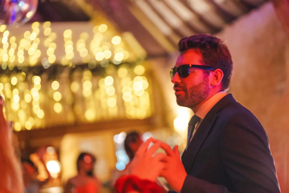 A groom wearing sunglasses and a dark suit enjoys the evening reception amidst a warm red glow and a sparkling bokeh background.