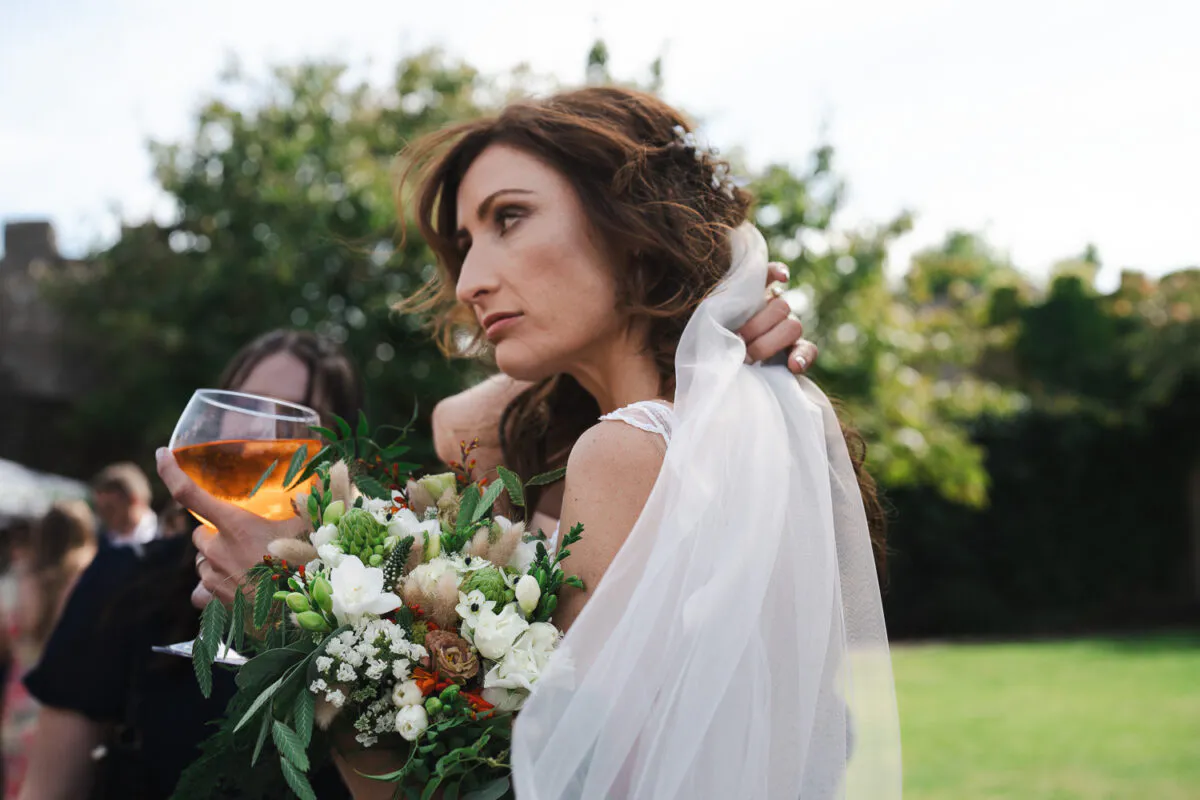 A bride with a half-up hairstyle and fingertip veil holding a lush hand-tied bouquet outdoors while holding a glass during the drinks reception.