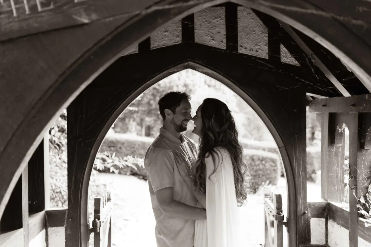 A newlywed couple shares a joyful nose-to-nose laugh while framed inside a traditional wooden church archway in a timeless black-and-white portrait.