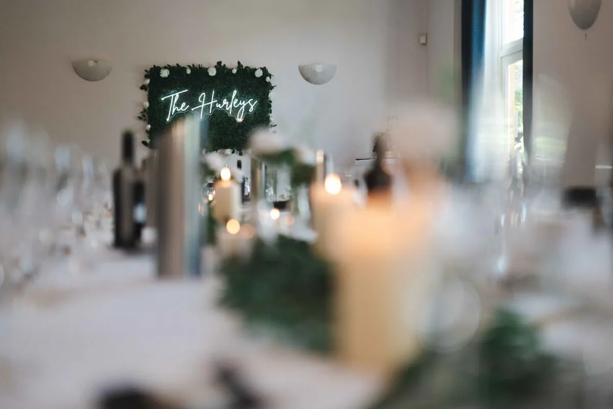 A close-up tablescape shot looking down a long banquet table featuring a greenery runner, glowing pillar candles, and a custom neon sign on a foliage wall in the background.