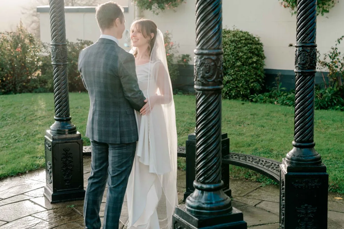 A bride and groom share a tender moment standing together outdoors under an ornate black bandstand during their wedding portrait session.