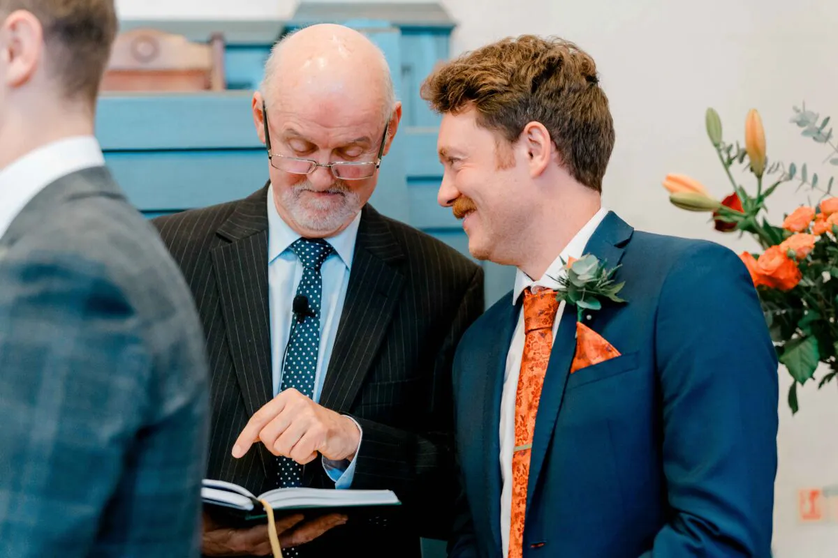 A smiling groom in a navy suit with an orange patterned tie looks down toward a celebrant who is pointing at a ceremony book.