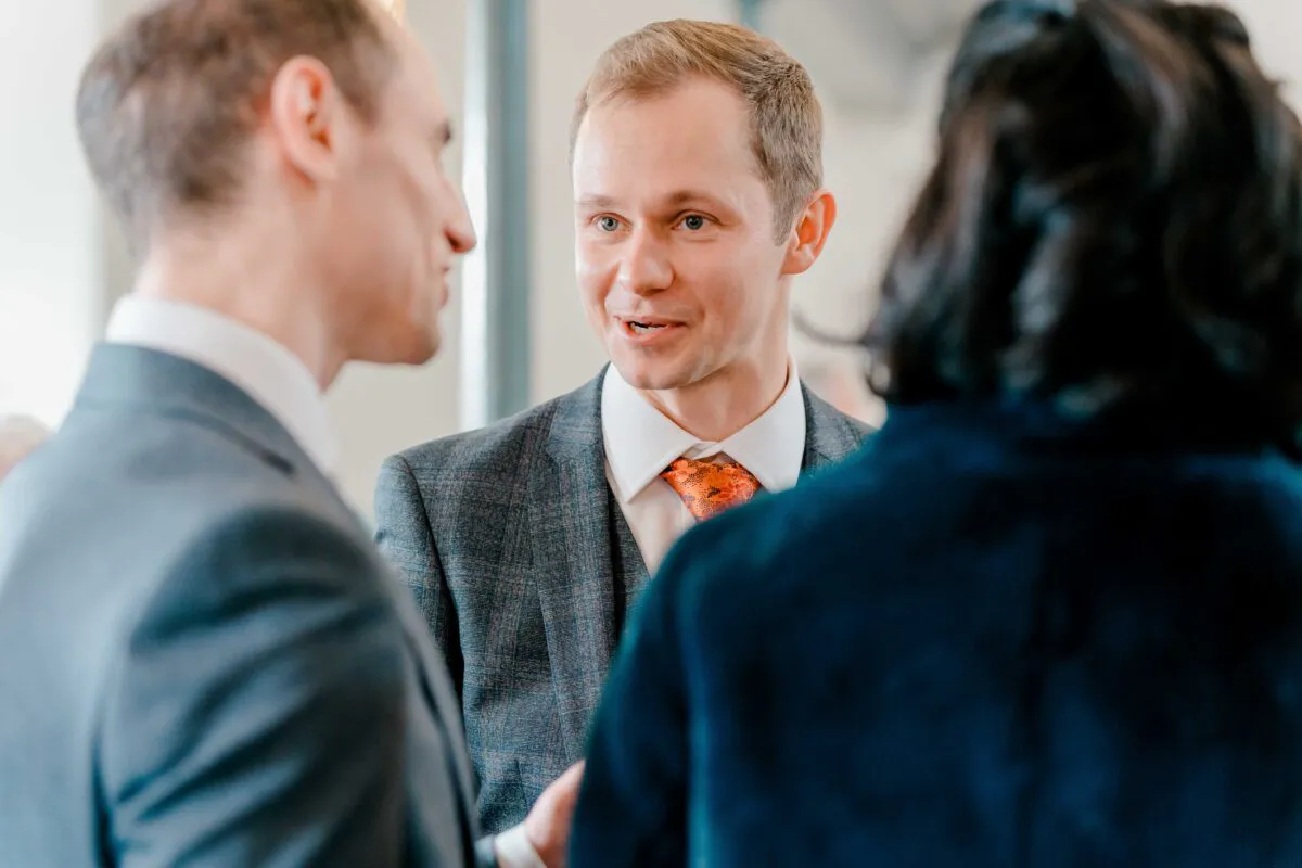 A guest in a grey patterned suit and orange tie chats with other wedding attendees during the drinks reception.