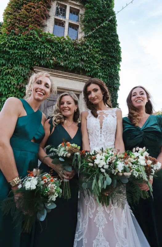 The bride and her three bridesmaids pose outside against an ivy-covered stone wall, holding lush bouquets with white florals and eucalyptus.