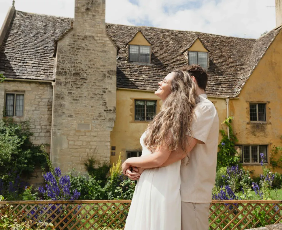A newlywed couple embraces lovingly outdoors in the sunshine with a charming historic stone manor house and vibrant garden flowers in the background.