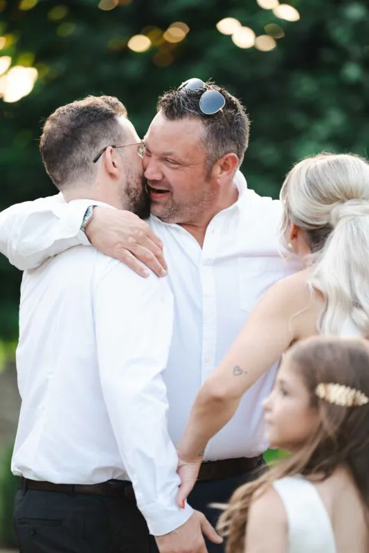 Two grooms share a joyful embrace outdoors while wearing white shirts, captured in candid light with guests nearby.
