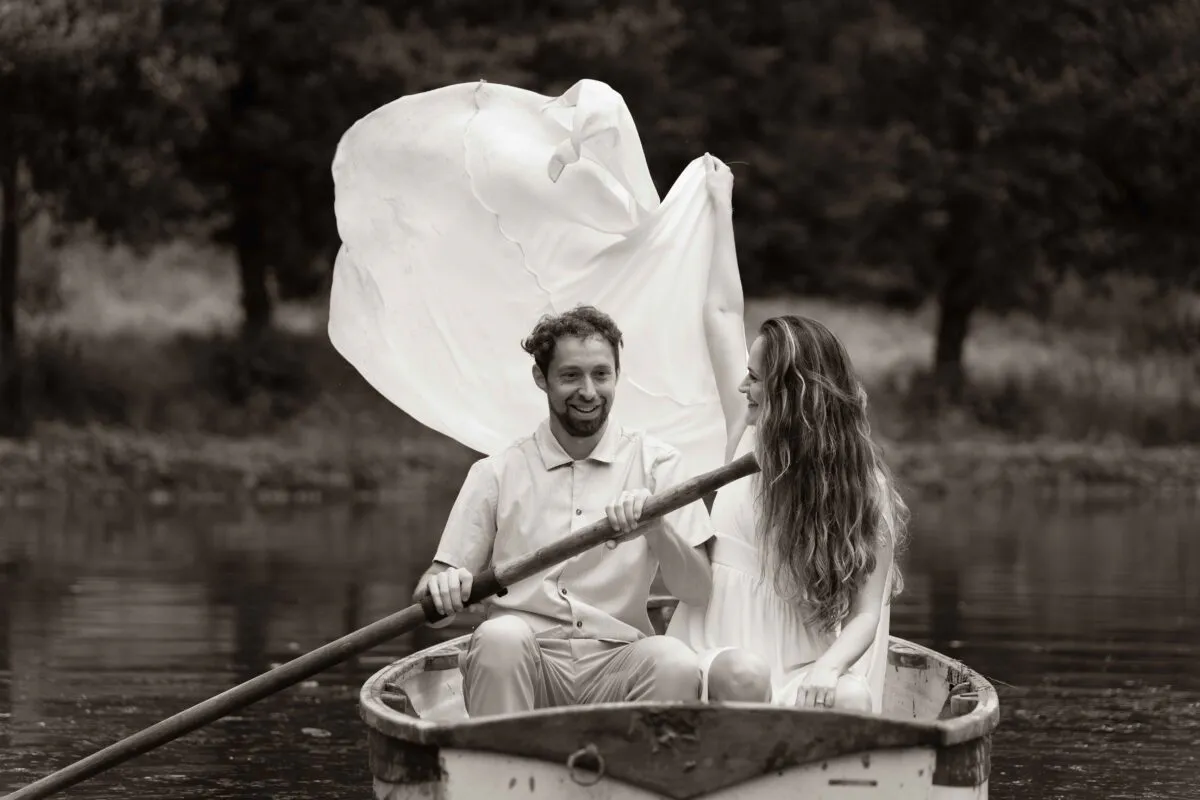 A newlywed couple laughing together in a small wooden rowing boat on a lake while fabric billows dramatically in the wind behind them.