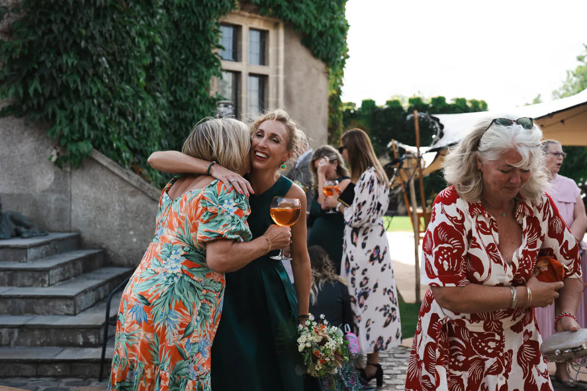 Guests enjoy drinks on a sunny afternoon in a stone courtyard, with two women embracing warmly and laughing while holding glasses.