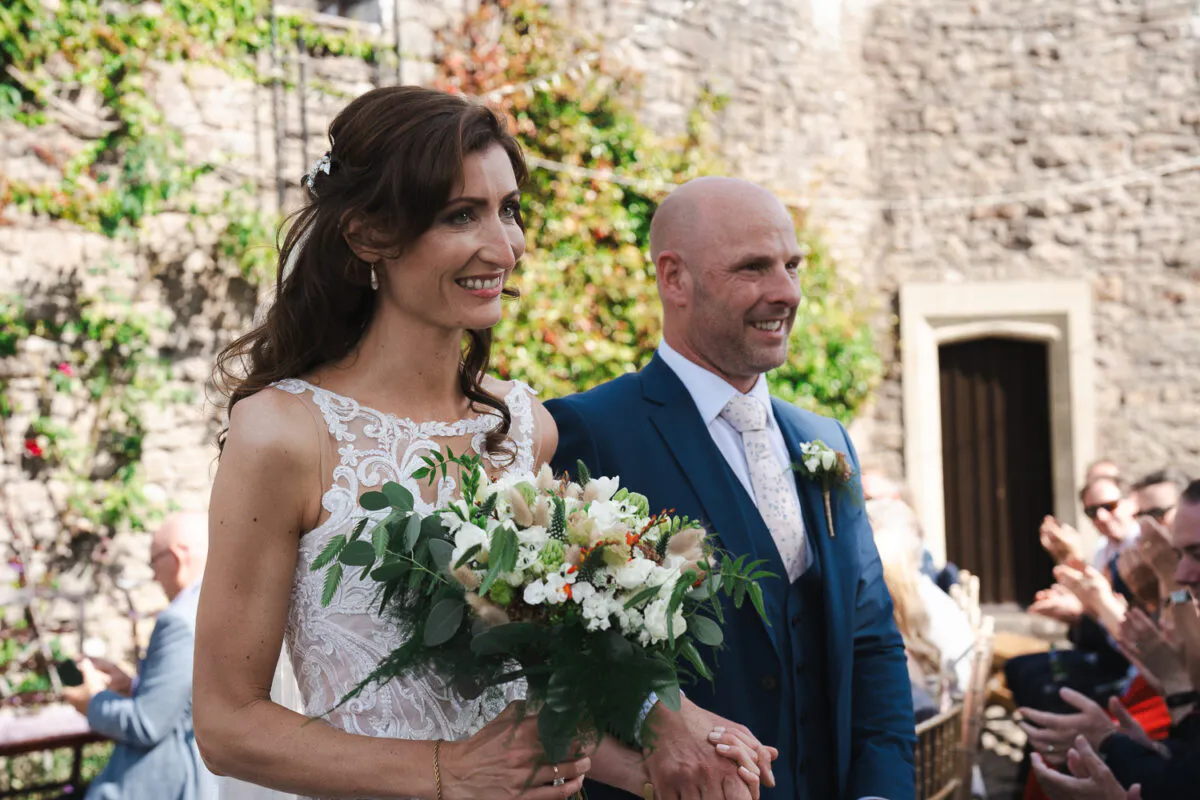 A smiling newlywed couple walks hand in hand past applauding guests after an outdoor ceremony against a historic stone wall.