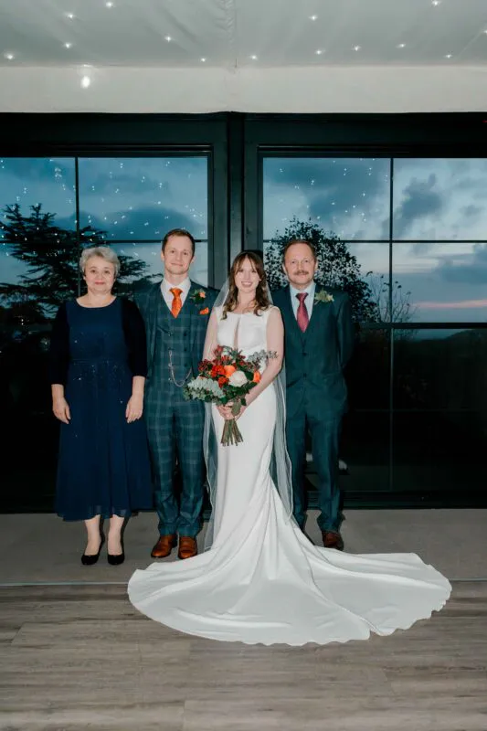 A newlywed couple poses proudly alongside the bride's parents against a large glass window at twilight, with the bride holding a vibrant bouquet.