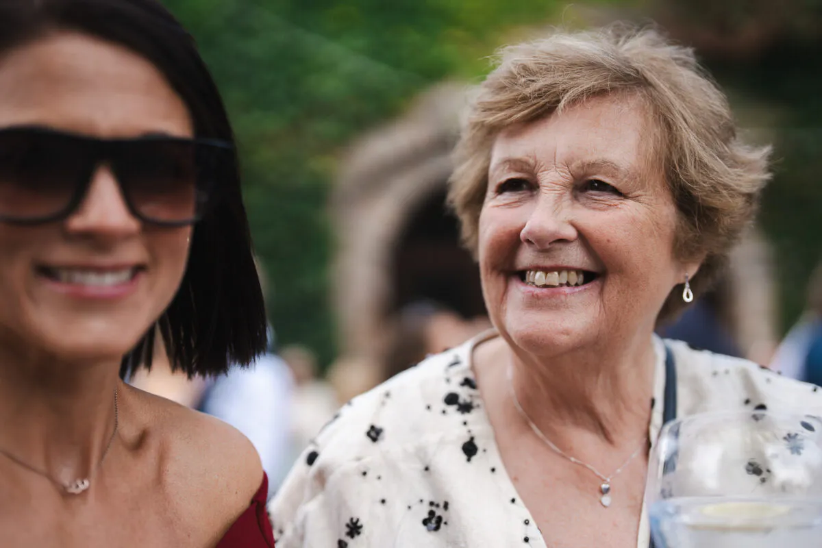 An older woman smiling joyfully during a candid wedding reception moment while a guest wearing sunglasses is partially visible in the foreground.