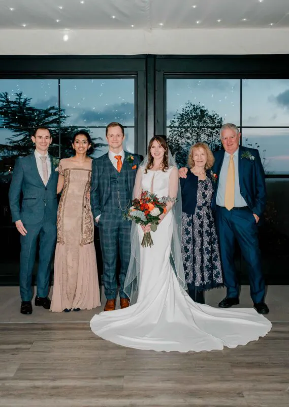 A newlywed couple posing with their wedding party and parents in front of large glass windows during an evening reception.