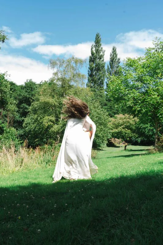 A newlywed couple is captured from behind running excitedly across a sunlit green lawn framed by tall trees under a clear blue sky.