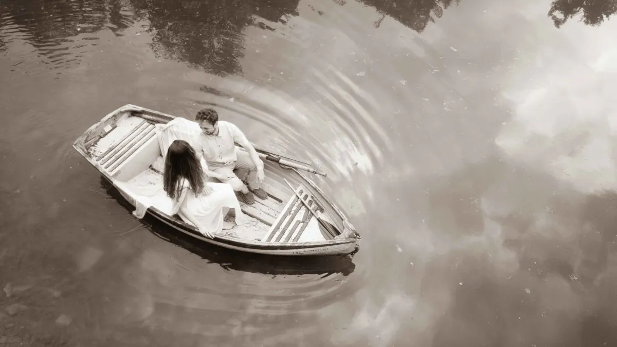 An aerial sepia-toned shot captures a newlywed couple sitting peacefully together in a wooden rowing boat on a calm lake.