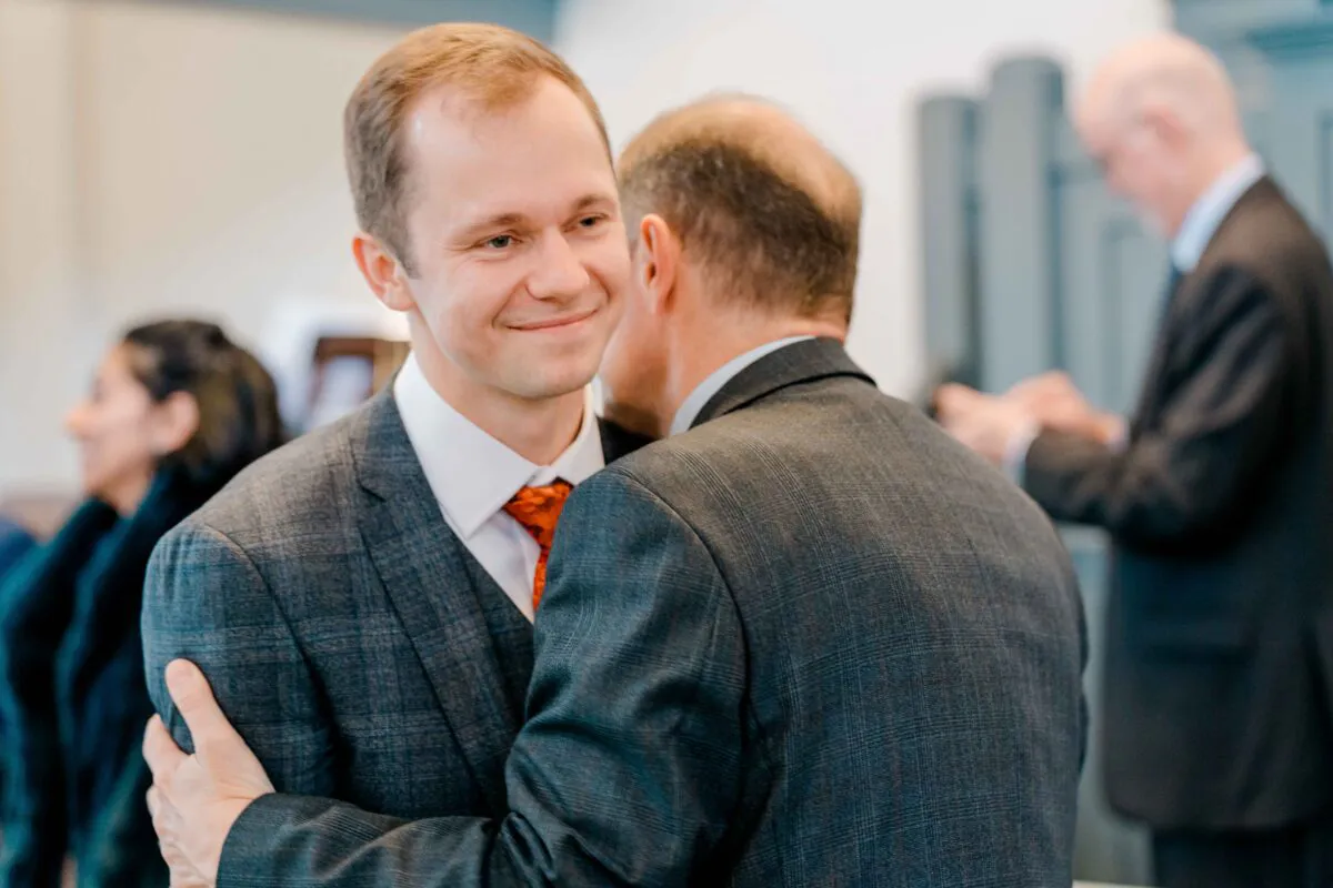 A smiling groom embraces a guest warmly during the wedding reception, wearing a tailored charcoal checked suit.