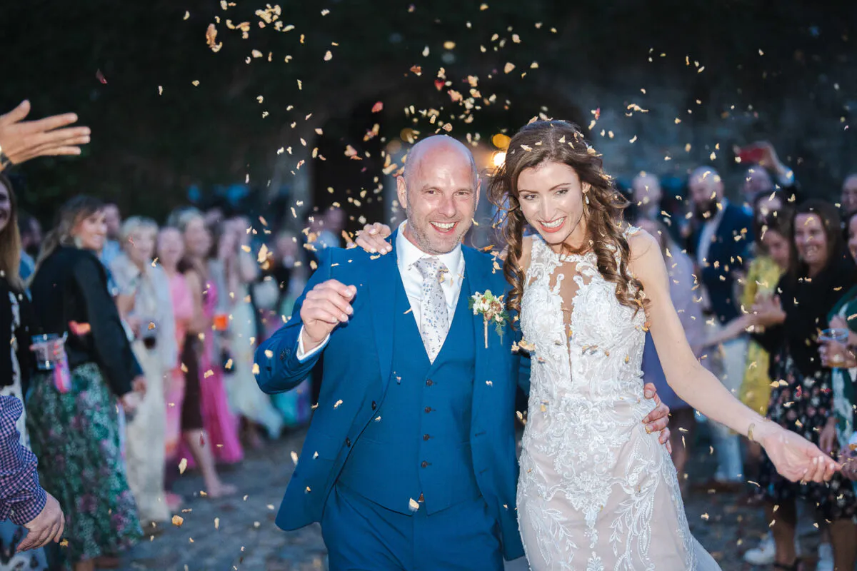 A joyful newlywed couple laugh as they walk through a shower of confetti thrown by cheering guests in a cobbled courtyard.