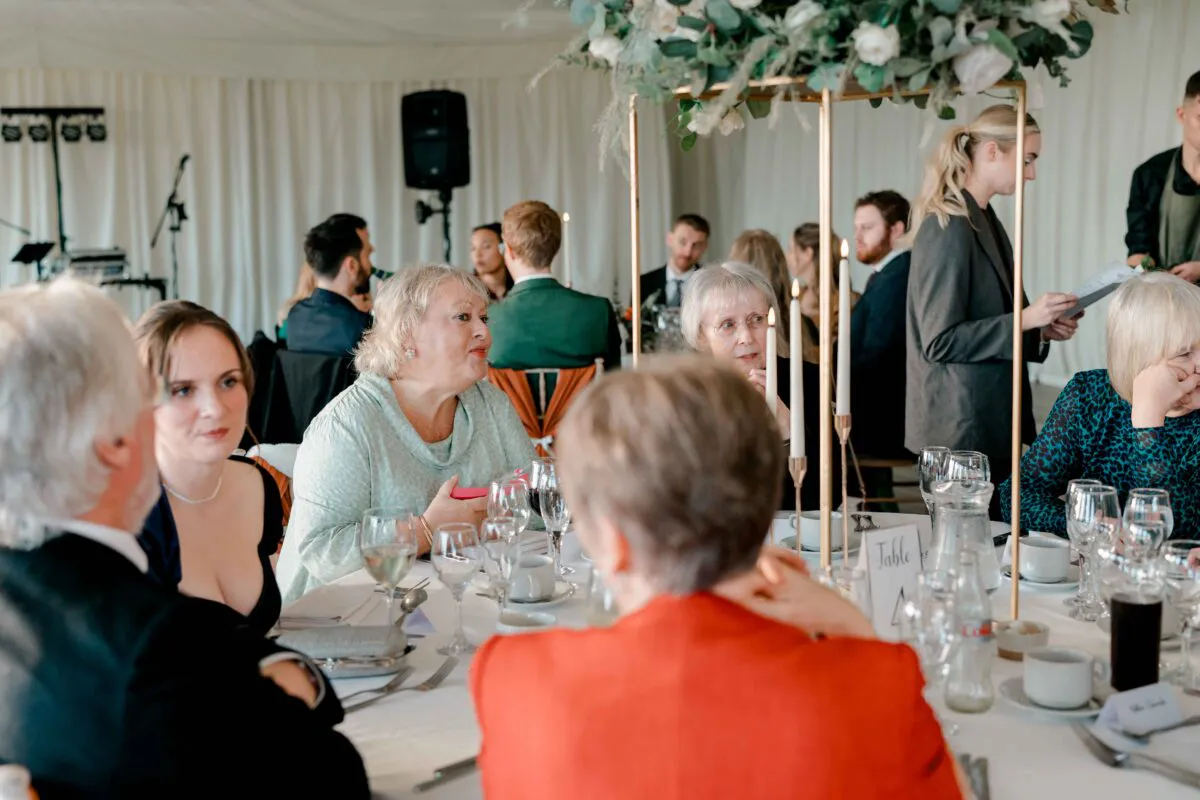 Wedding guests enjoy conversation and drinks seated at tables adorned with taper candles during a wedding breakfast inside a bright marquee.