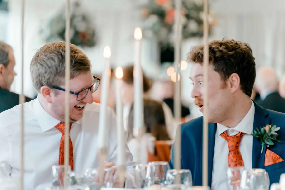 Two grooms in matching patterned ties and white shirts share a joyful, laughing moment during the wedding breakfast, framed by tall candlesticks.