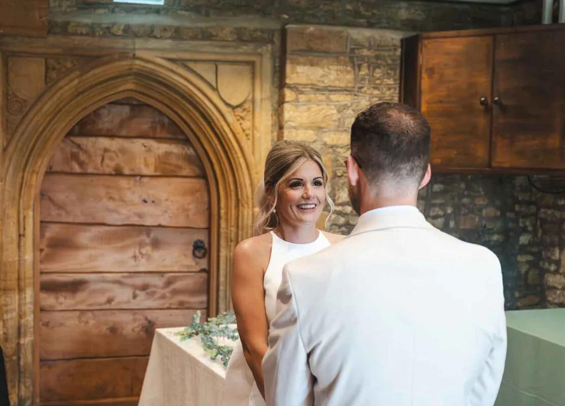 A smiling bride looks lovingly at the groom during an intimate ceremony inside a historic venue with a prominent stone archway.