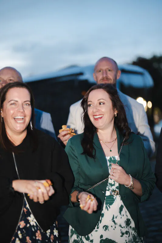 Wedding guests laughing joyfully while holding small items outdoors on a cobbled courtyard during an evening reception.