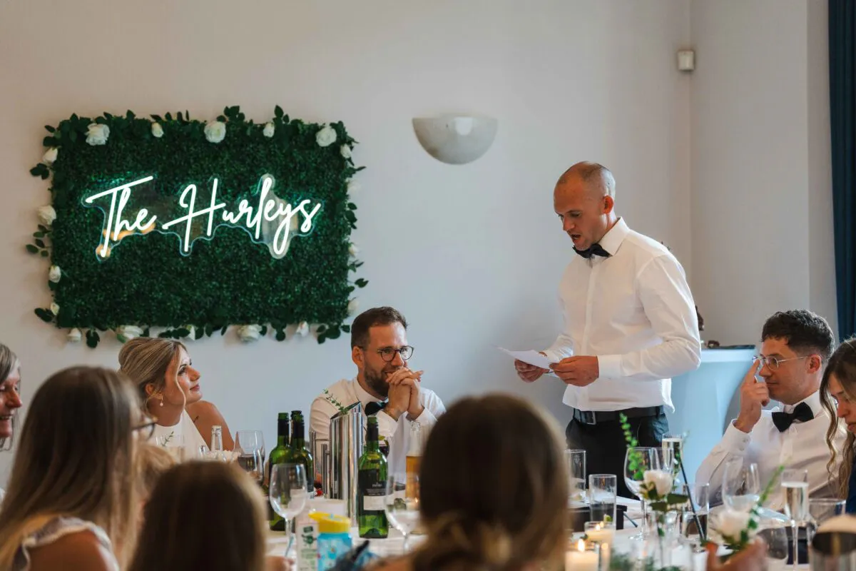 A groom delivers a speech while holding paper notes at the top table, set against a white wall featuring a glowing neon sign mounted on a lush greenery backdrop.