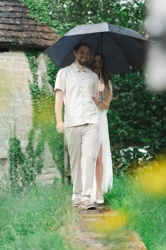 A smiling bride and groom walk together outdoors under a black umbrella during a rainy wedding day, with an ivy-covered stone wall in the background.