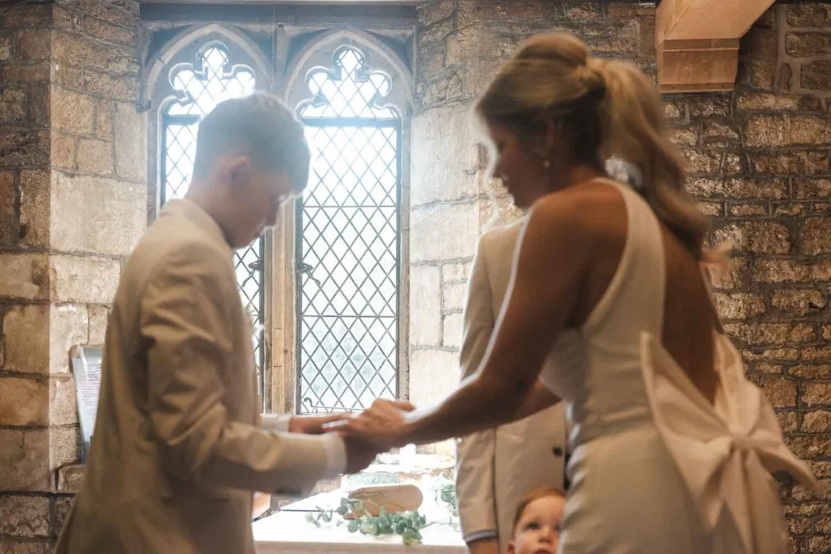 A bride with an oversized bow on the back of her dress and a high ponytail holds hands indoors near a historic leaded window during a wedding ceremony.