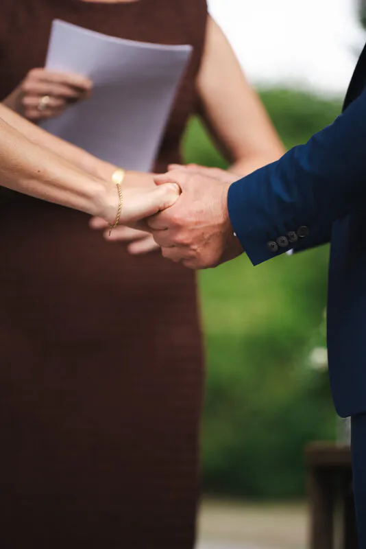 A close-up shot capturing a couple holding hands during an outdoor wedding ceremony while a celebrant stands nearby holding papers.