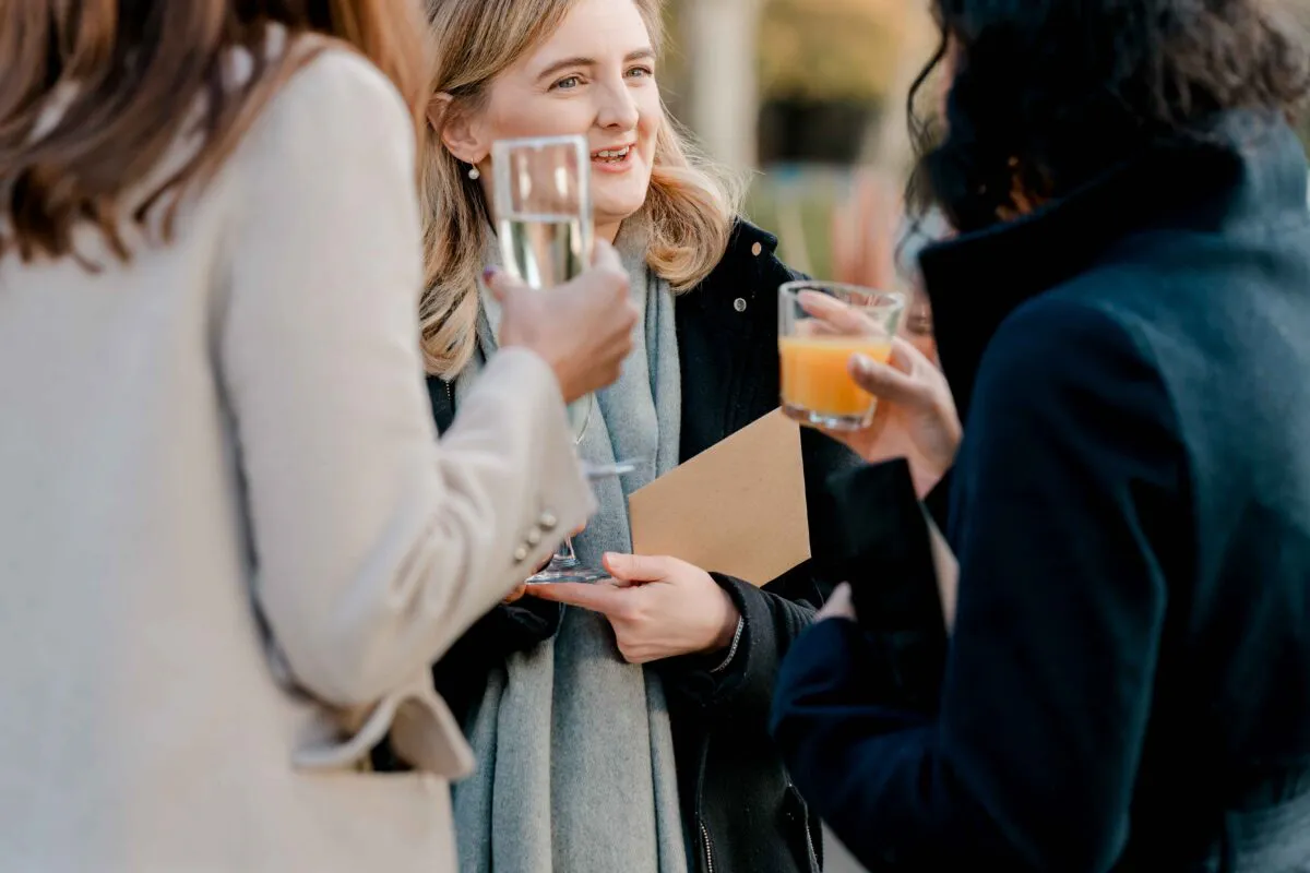 Three wedding guests chat and share a drink outdoors during the daytime drinks reception.
