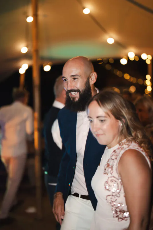 A joyful couple smiles happily together under a tent illuminated by glowing fairy lights during an evening wedding reception.