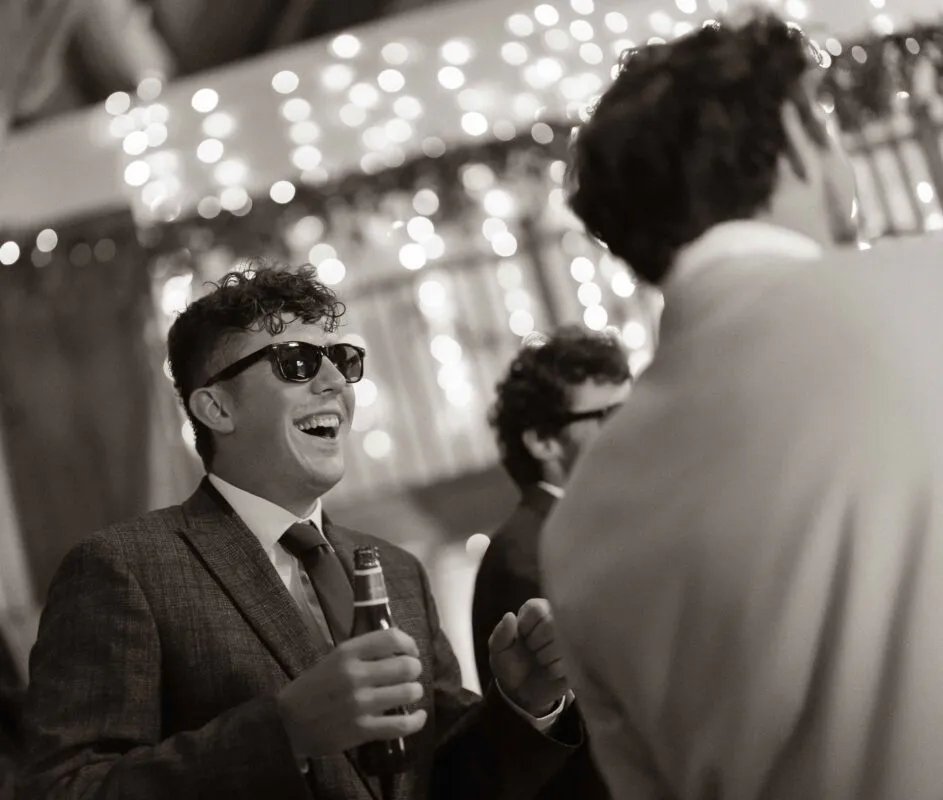 A guest wearing sunglasses and a tweed suit laughs joyfully while holding a beer bottle amid a twinkling fairy light backdrop during the wedding reception.