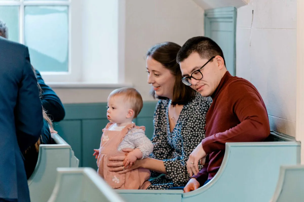 A family with a young baby sitting together in light blue church pews attending a wedding ceremony.