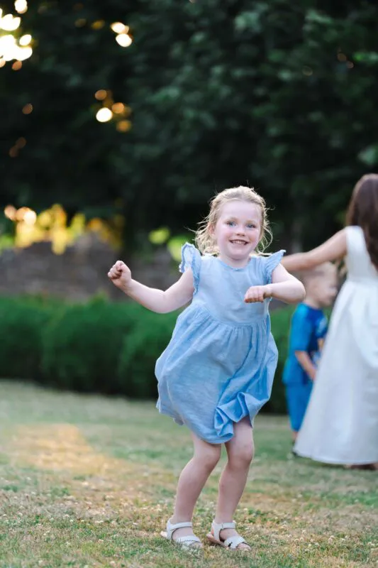 A young flower girl in a light blue dress dances joyfully on a lawn during an outdoor wedding reception.