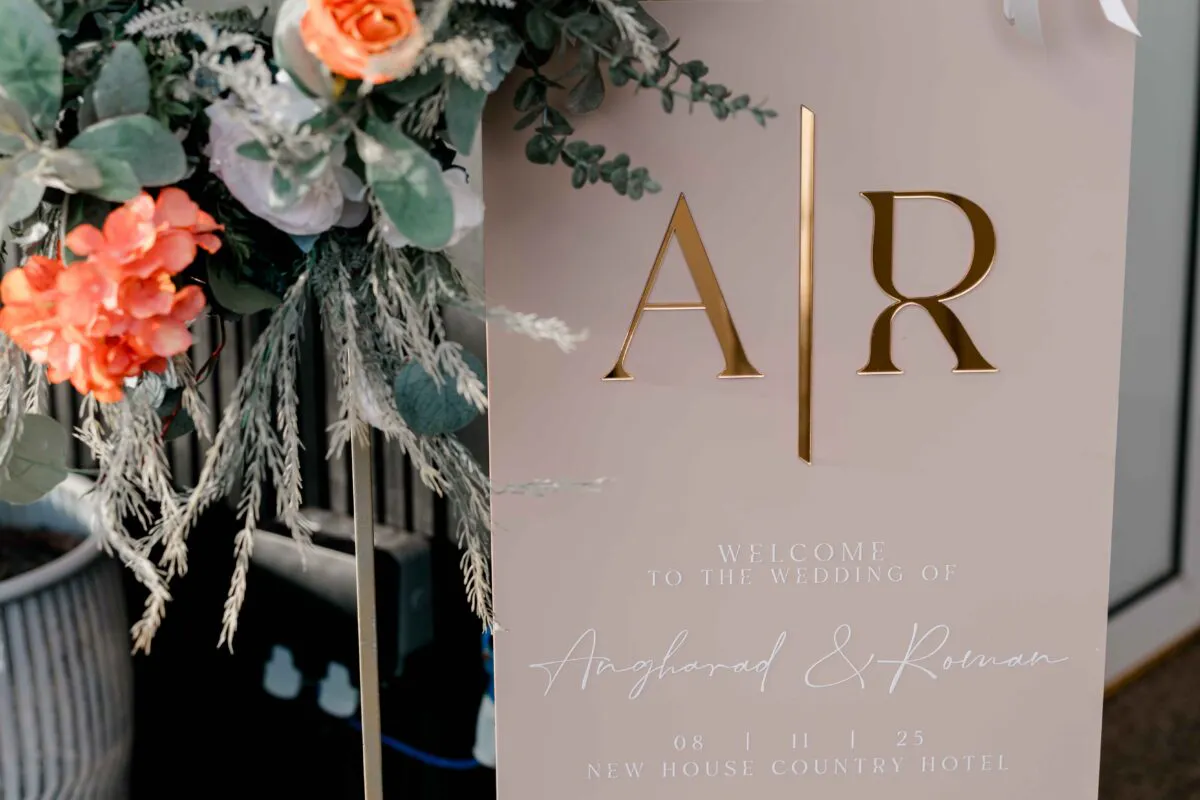 An elegant modern wedding welcome sign featuring gold acrylic monogram initials and script lettering on a pale backing, adorned with vibrant coral and white floral arrangements.