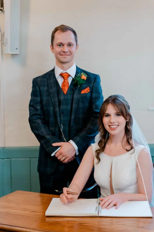 A smiling bride sits at a wooden table signing the wedding register, wearing a white dress with a keyhole neckline and a veil, while the groom stands behind her in a checked suit and orange tie.