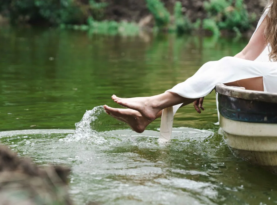 A barefoot bride dips her feet into a tranquil river from the edge of a wooden rowing boat, kicking up water splashes.