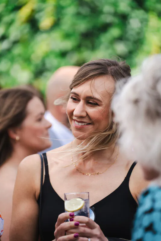 A guest smiles and holds a drink with a lemon slice during an outdoor wedding drinks reception.
