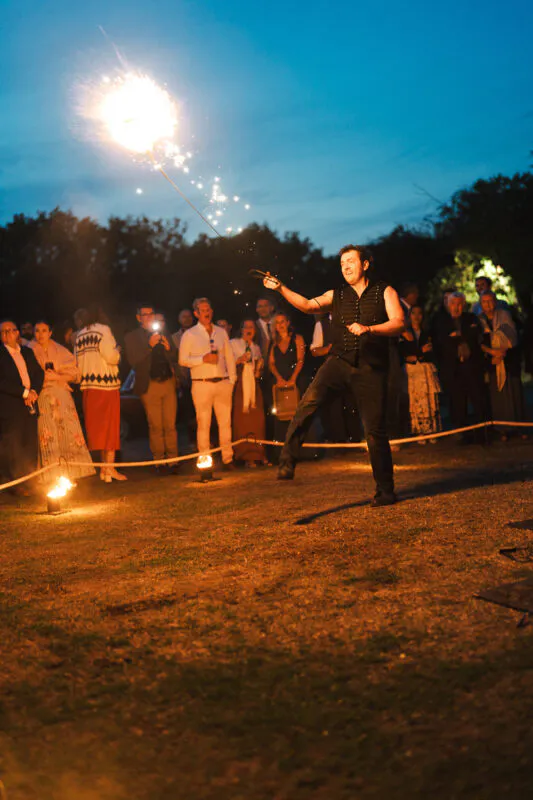 A fire performer swings a glowing spark on a tether during the evening reception as wedding guests watch enthusiastically under a blue hour sky.