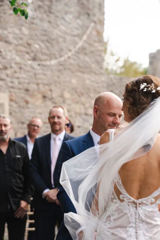 An emotional groom looks at his bride during an outdoor ceremony in front of a stone wall, with a veil draped softly in the foreground.