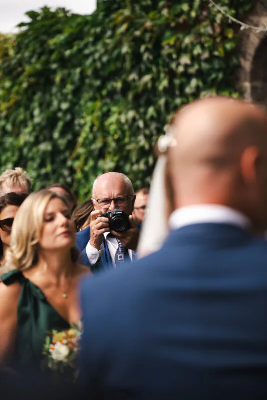 An older guest wearing glasses takes a photograph with a digital camera among a crowd of onlookers in front of an ivy covered wall.