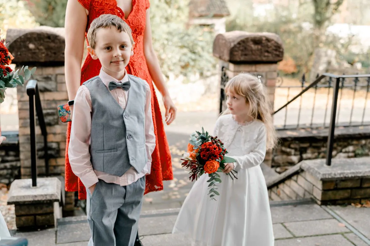 A young page boy in a grey waistcoat and bow tie poses next to a flower girl holding a vibrant red and orange bouquet outdoors.