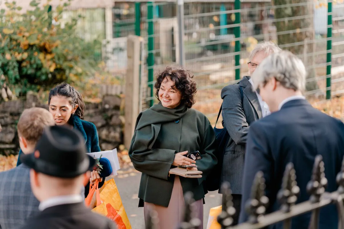 Wedding guests mingling and smiling outdoors during an autumn celebration near a metal fence.