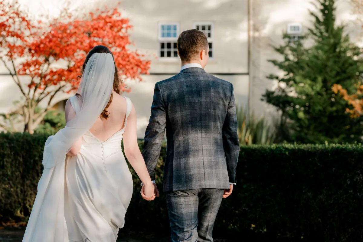 Newlyweds walk hand in hand away from the camera in a scenic autumn garden with vibrant red foliage and a stately home in the background.