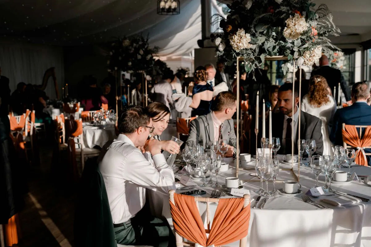 Guests enjoy a wedding breakfast seated at round tables inside a sunlit marquee decorated with tall floral centrepieces and warm candles.
