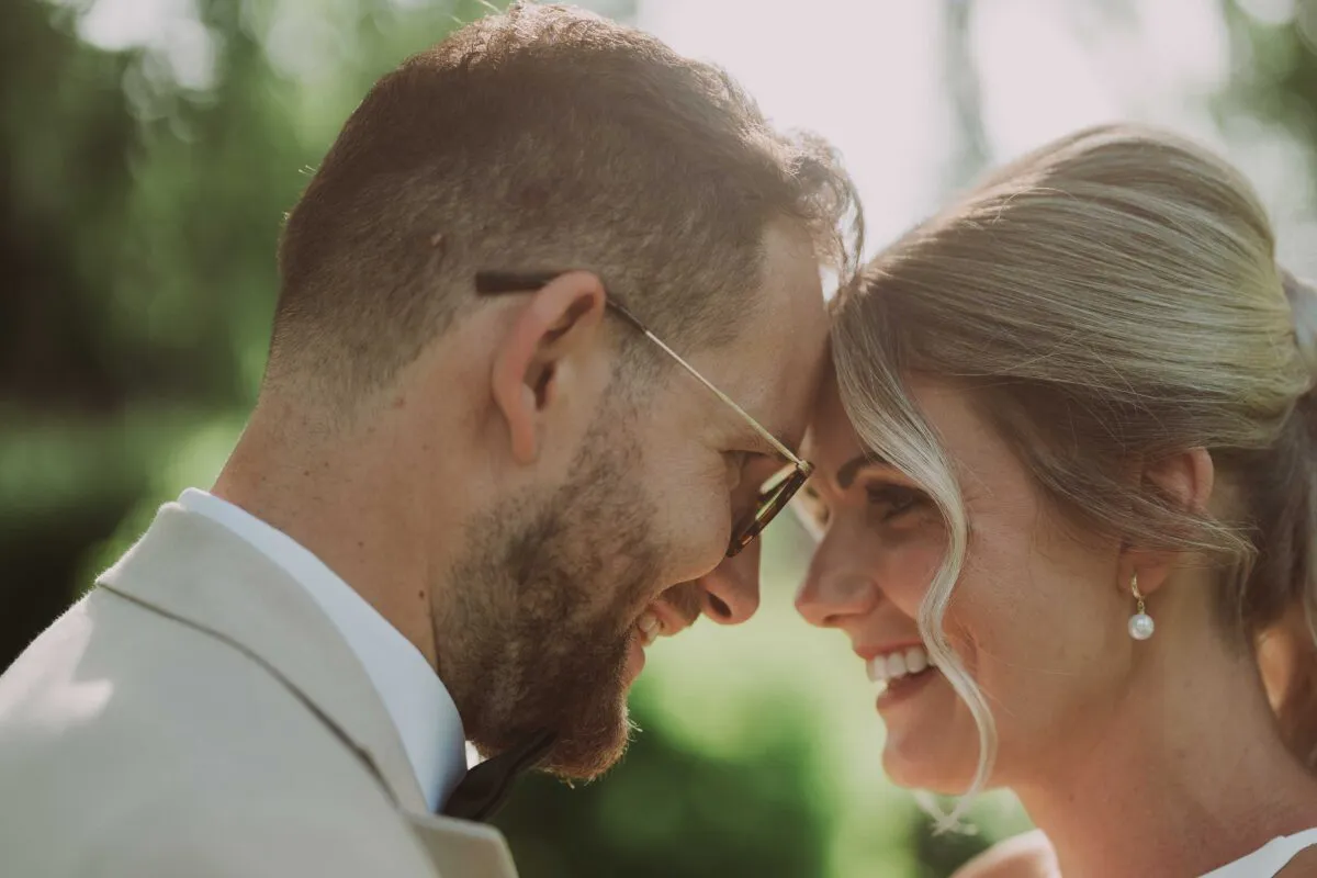 A close-up profile portrait of a smiling bride and groom pressing their foreheads together outdoors in warm backlighting.