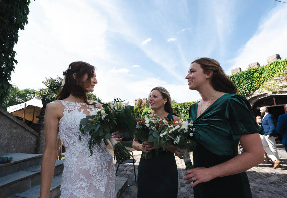 A low-angle outdoor shot of a bride in a lace gown holding a bouquet while chatting and laughing with two bridesmaids in emerald green velvet dresses against a sunny castle backdrop.