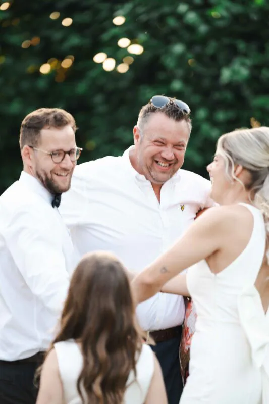 A smiling couple and wedding guests share a joyful moment outdoors surrounded by lush green foliage and dappled light.